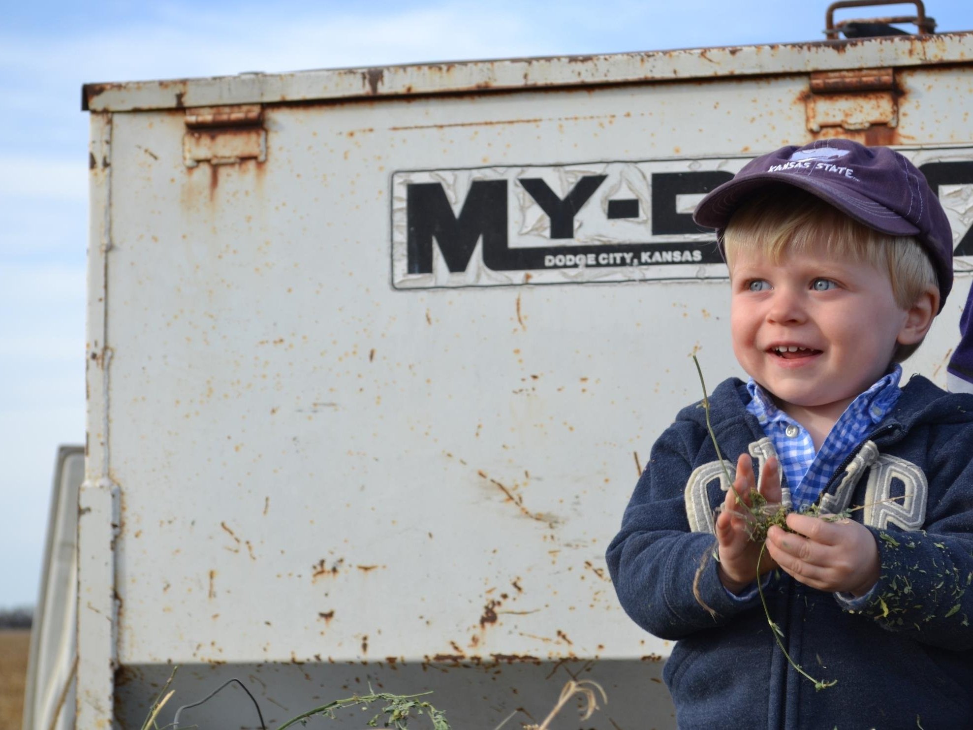 Young boy smiling while holding grass and standing in front of a rusty agricultural trailer, with a blue sky in the background. The child is dressed in a blue hoodie and a Kansas State cap, embodying a cheerful moment in a rural setting.